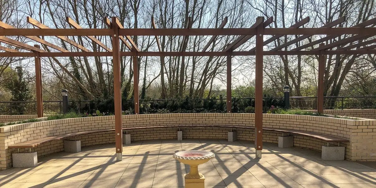 Outdoor dark wood pergola over curved wooden benches against curved wall and bird bath in the foreground