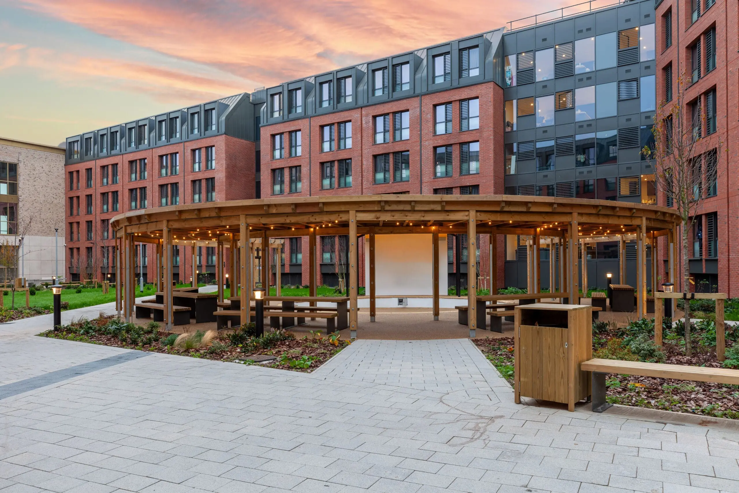 Modern building, with the sun setting in the background. Wooden canopy with benches and fairy lights.