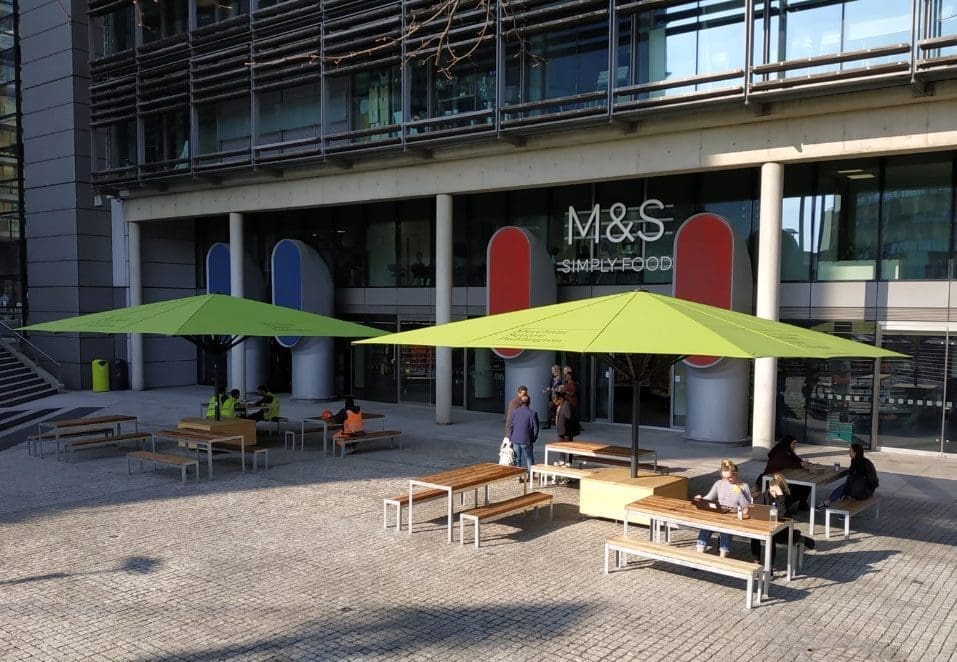 Exterior of M&S building with wooden and metal picnic benches and tables under large square green parasols