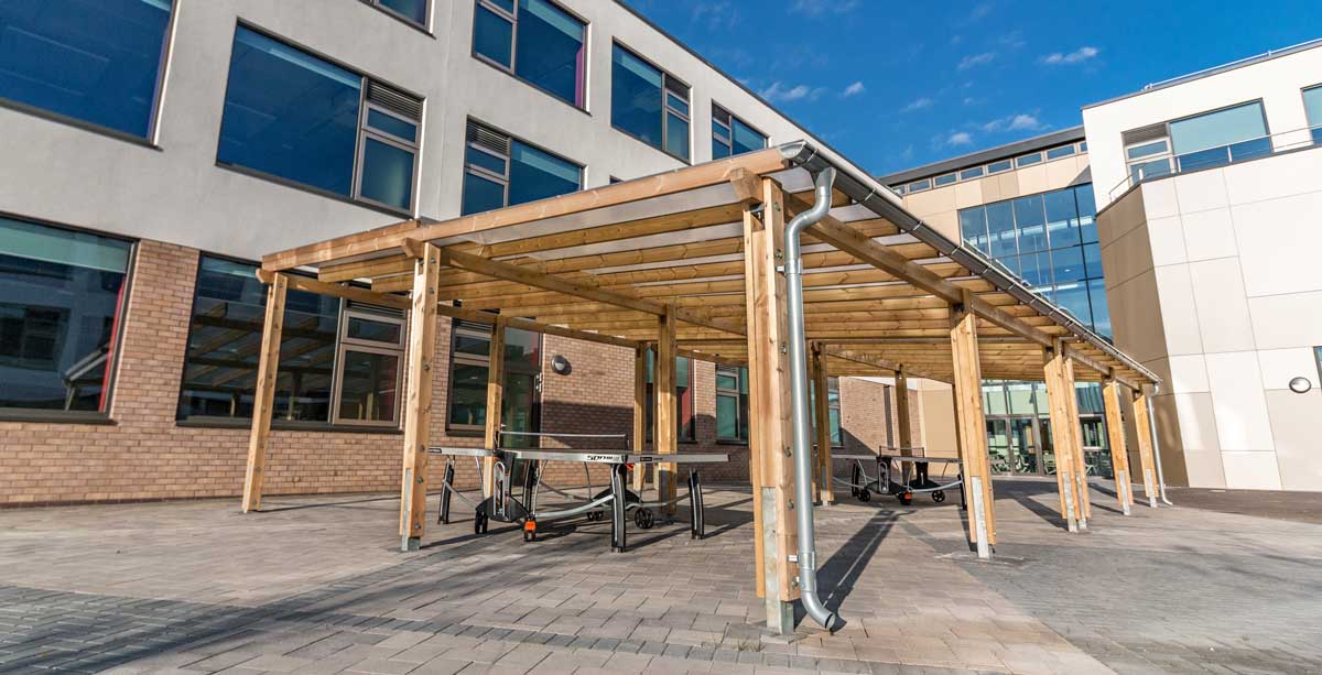 Large outdoor wooden pergola covering tablet tennis tables in school courtyard
