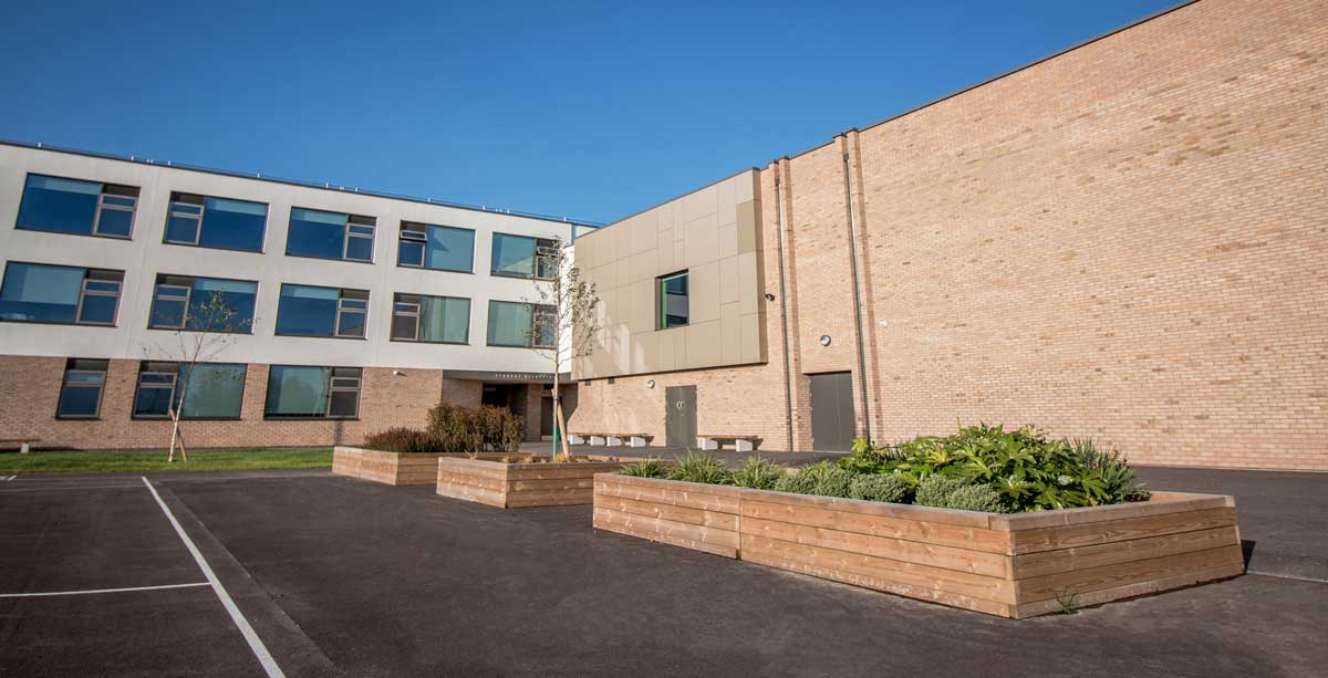 Row of large wooden raised planters alternating between bushes and trees planted inside infront of school