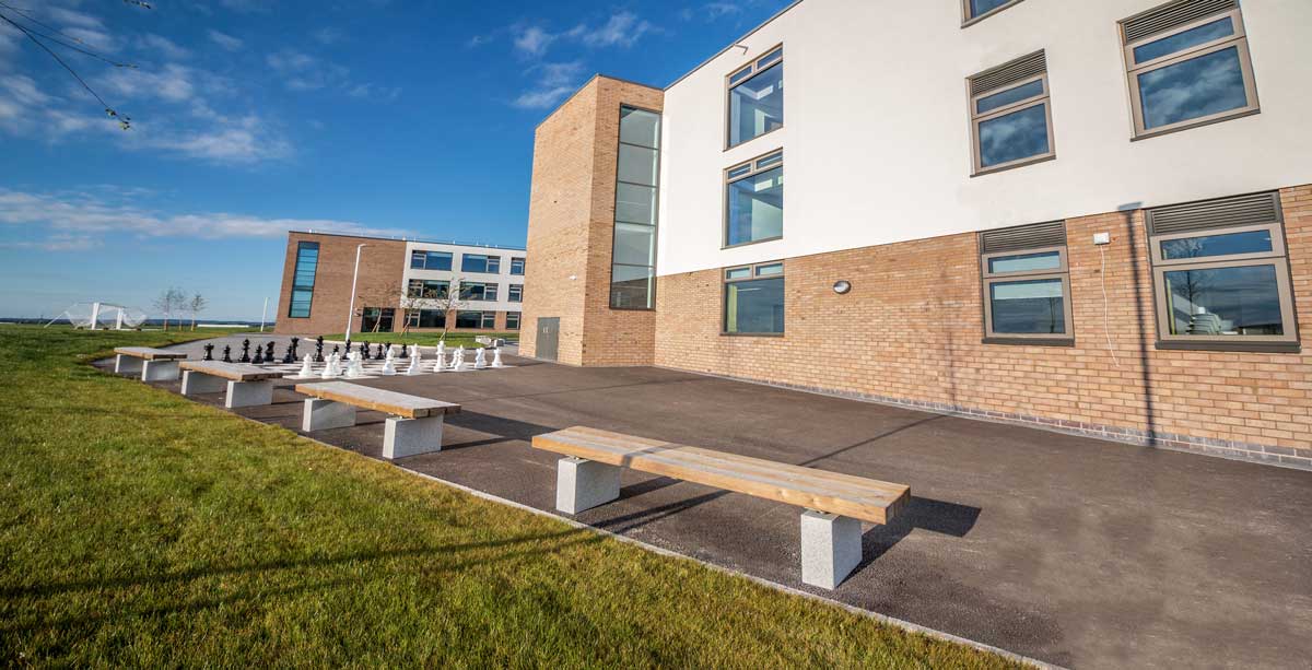 Row of wooden benches with concrete plinth legs on school playground next to large outdoor chess set infront of school building