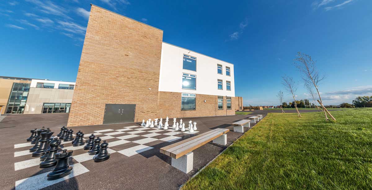 Row of wooden benches with concrete plinth legs on school playground next to large outdoor chess set infront of school building