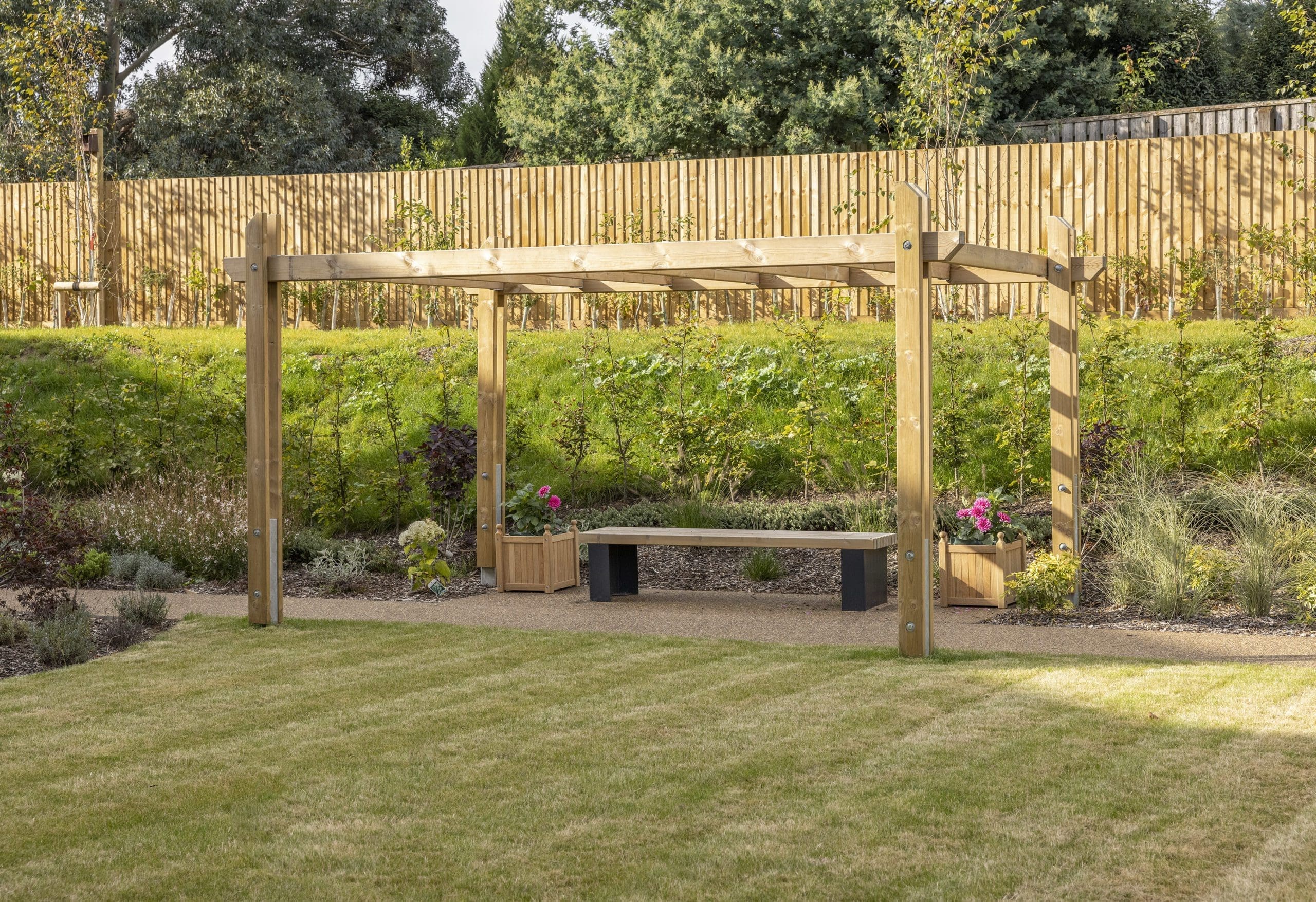 Outdoor wooden pergola canopy above wooden bench with black plinth legs and planters left and right