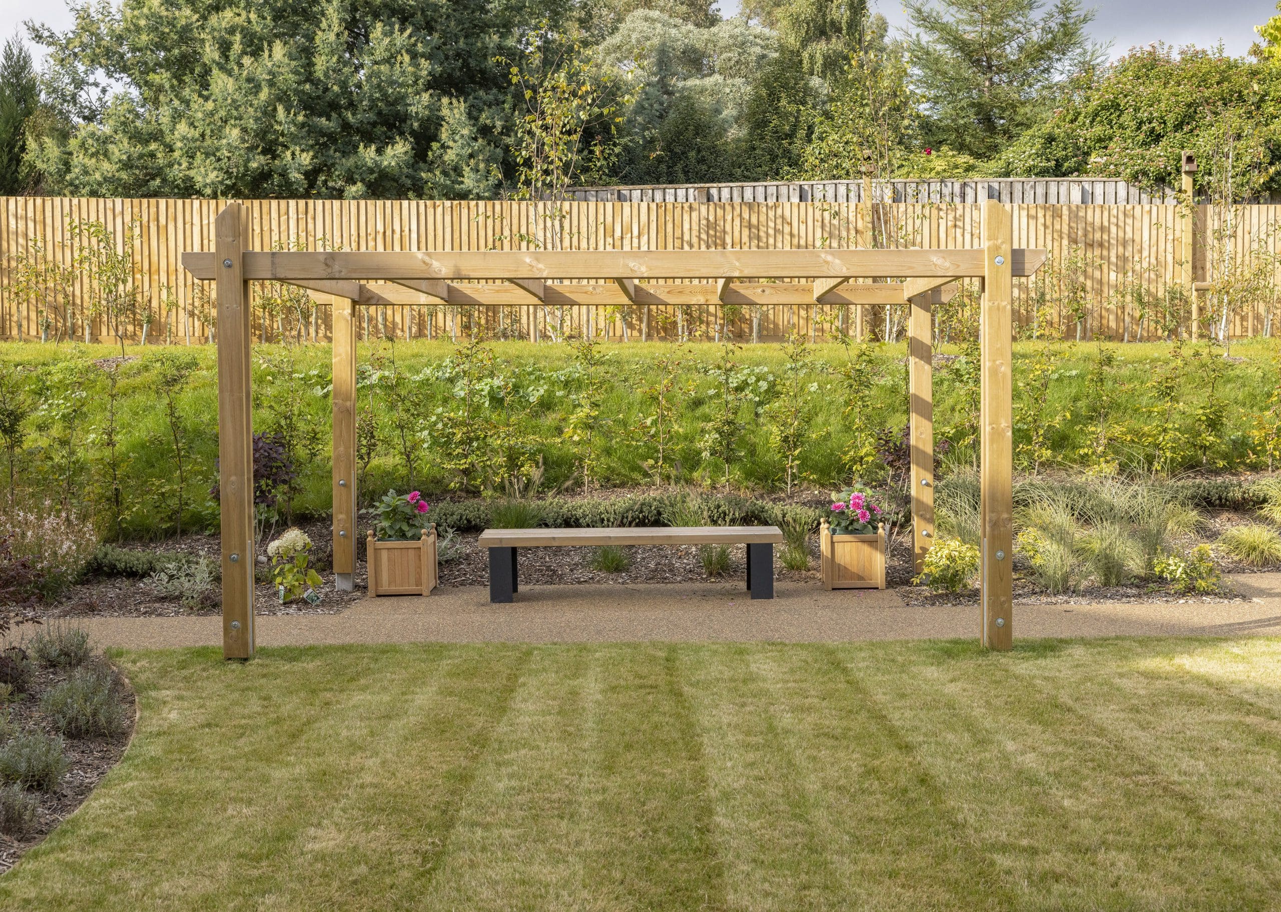Outdoor wooden pergola canopy above wooden bench with black plinth legs and planters left and right