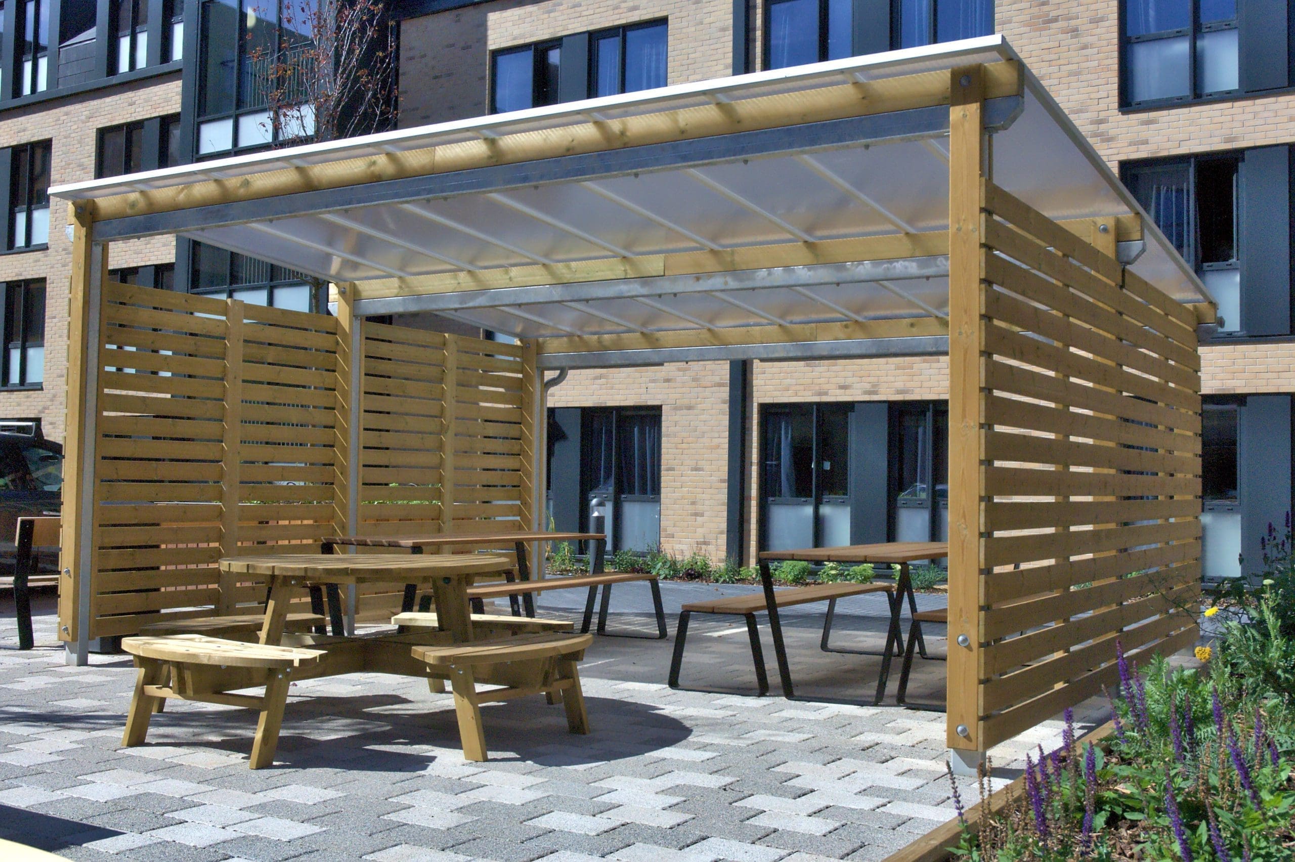 Outdoor canopy with wooden sides and see through plastic top covering a variety of wooden picnic tables and benches