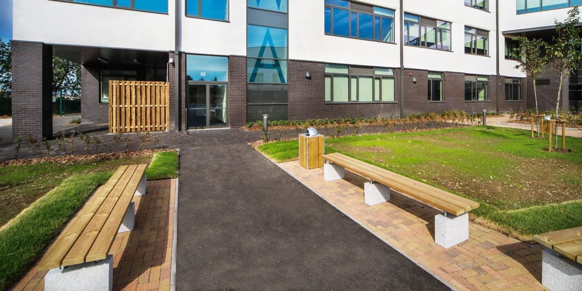 exterior shot of the school showing rows of wooden benches with concrete plinth legs surrounding greenery areas