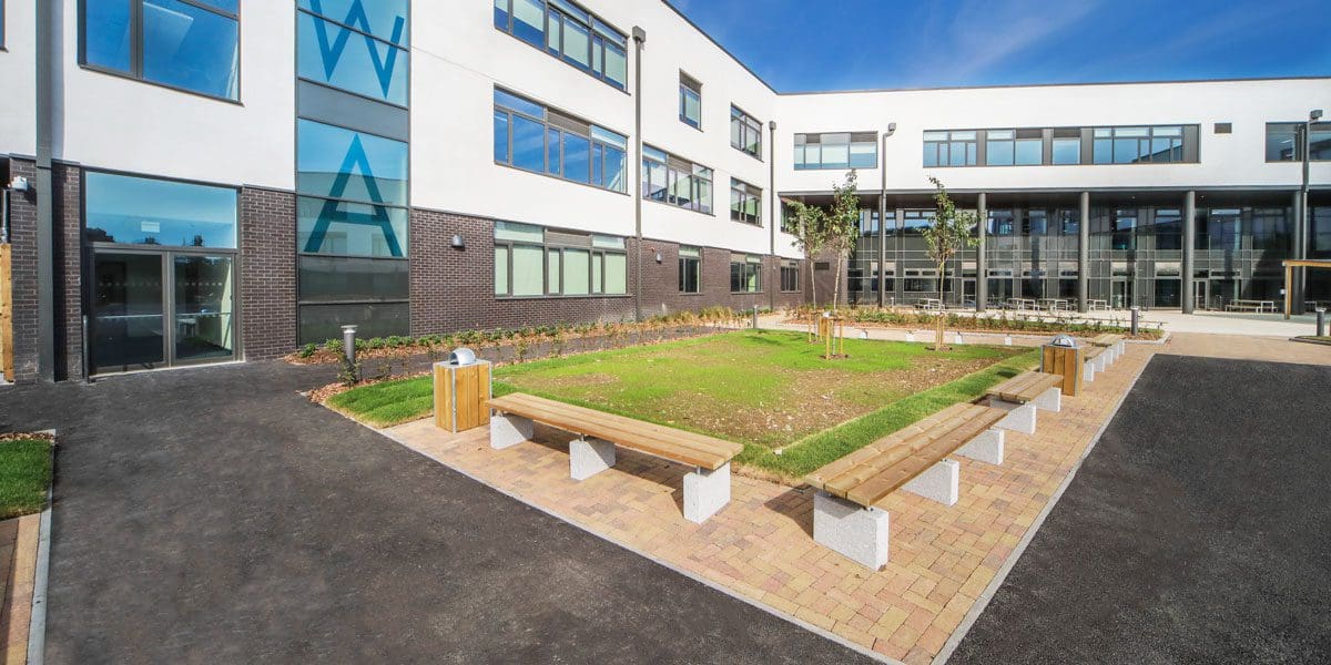 exterior shot of the school showing rows of wooden benches with concrete plinth legs surrounding greenery areas
