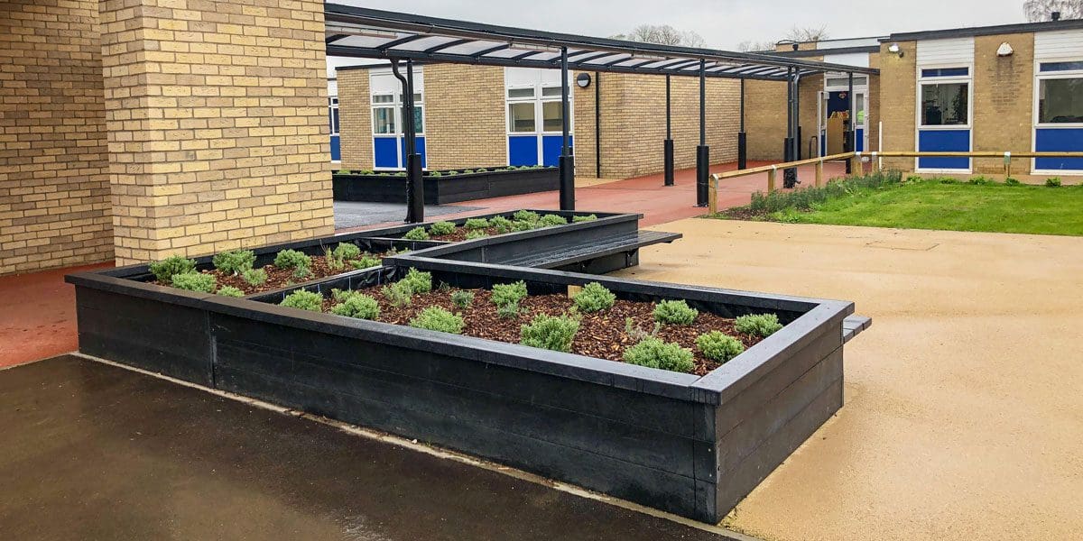 External black metal canopy with see through roof connecting two buildings with black wooden raised planters and attached benches in foreground