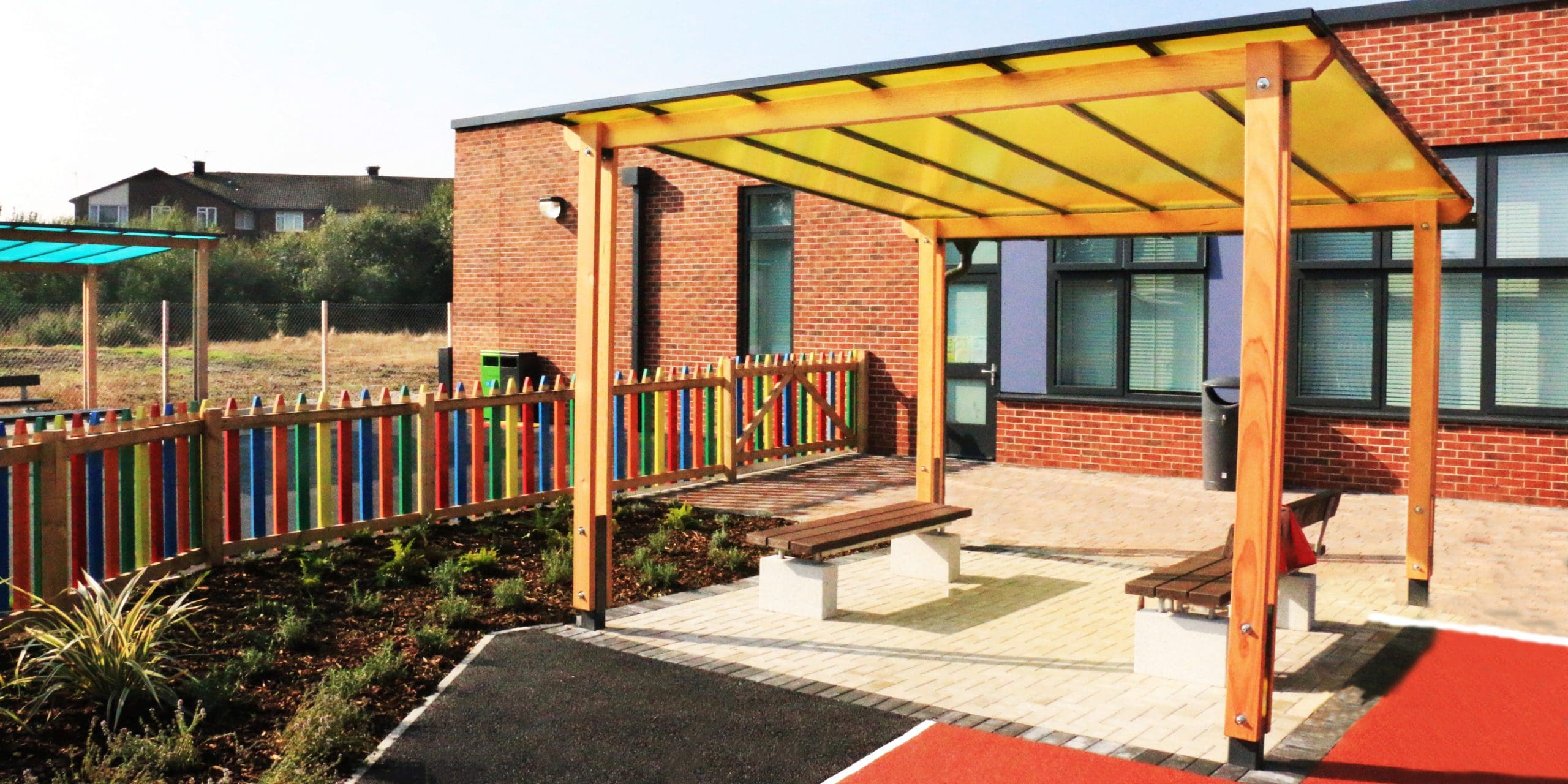 External pergola with yellow tinted canopy colour above dark brown benches with concrete plinth legs