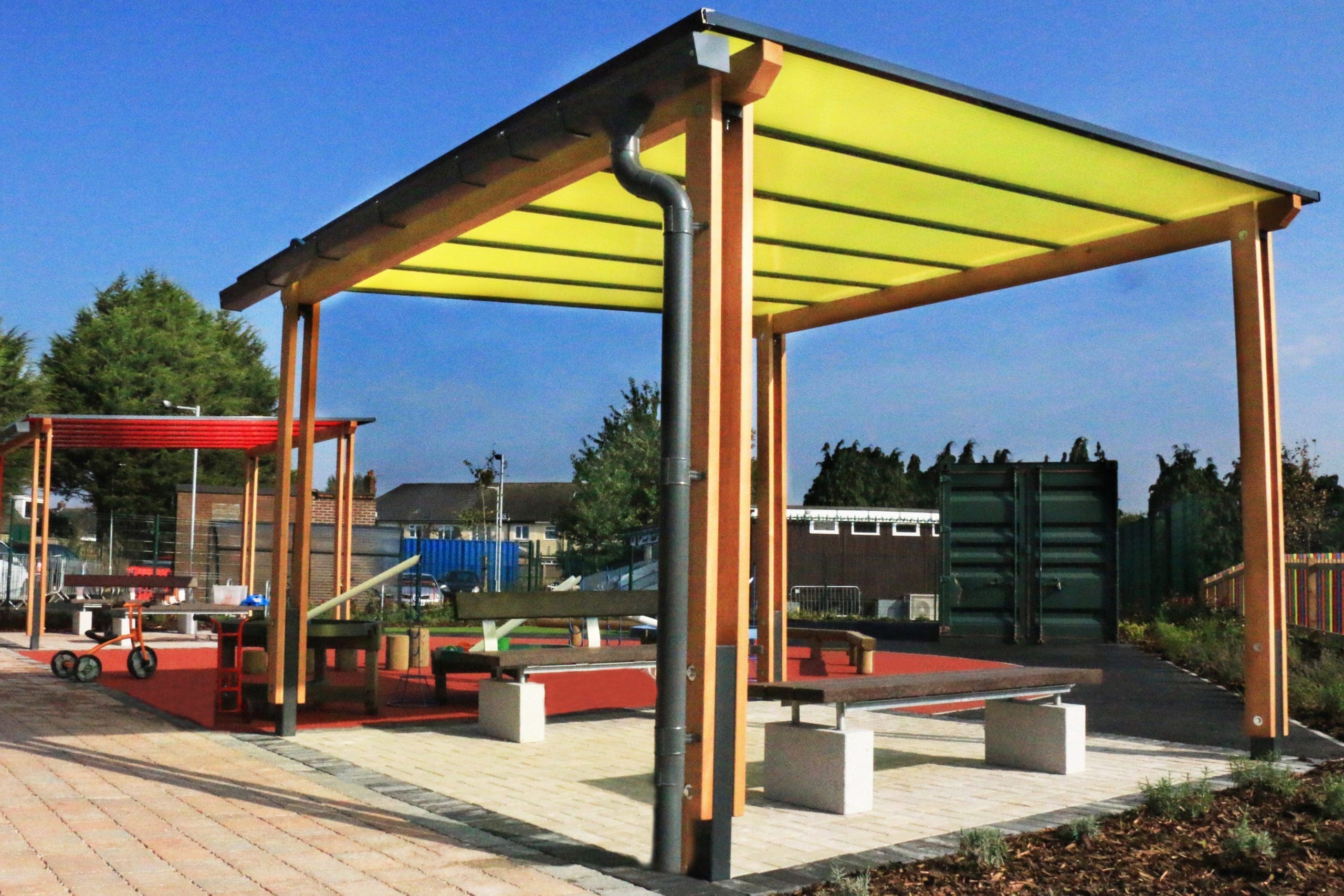 External pergola with yellow tinted canopy colour above dark brown benches with concrete plinth legs