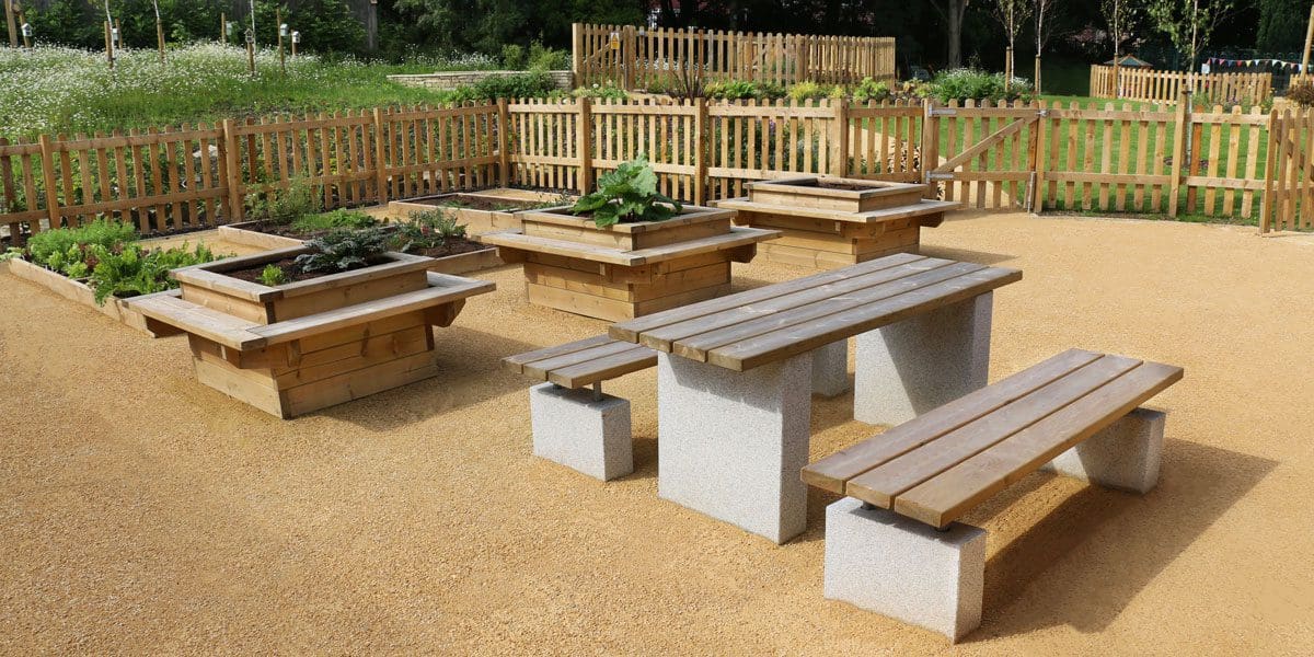 Rows of long raised plant beds and taller square planters with attached seating with wooden and concrete table and benches infront