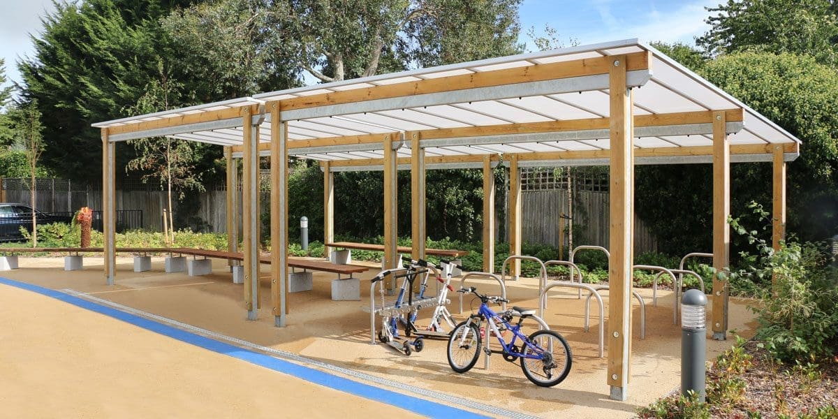 Large pergola with canopy covering bicycle and scooter parking racks and wooden benches with concrete plinth legs