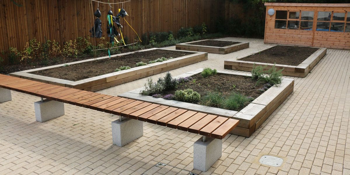 Outdoor wooden bench with concrete plinth legs infront of four square small raised plant beds