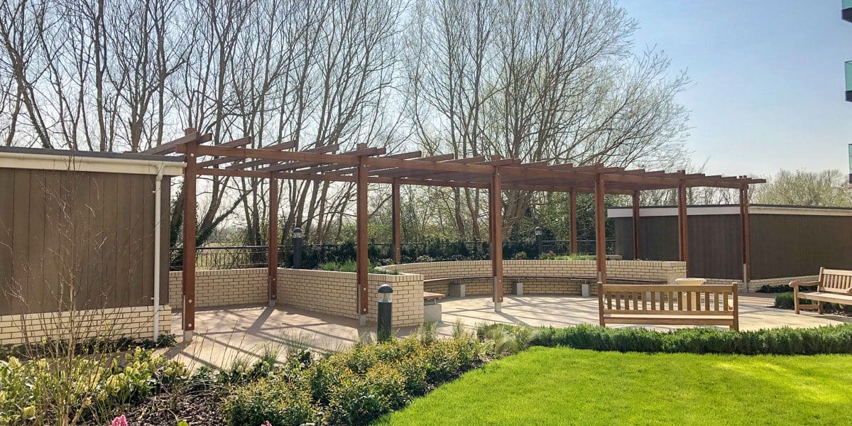 Outdoor dark wood pergola over curved wooden benches against curved wall and bird bath in the foreground