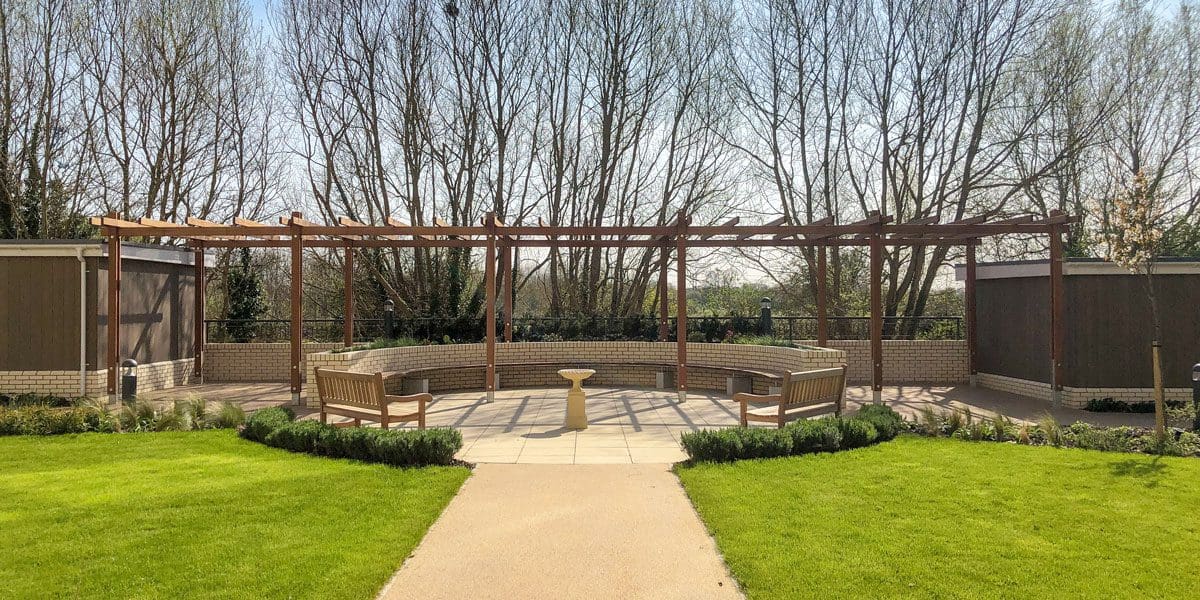 Outdoor dark wood pergola over curved wooden benches against curved wall and bird bath in the foreground
