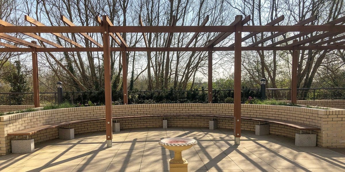 Outdoor dark wood pergola over curved wooden benches against curved wall and bird bath in the foreground