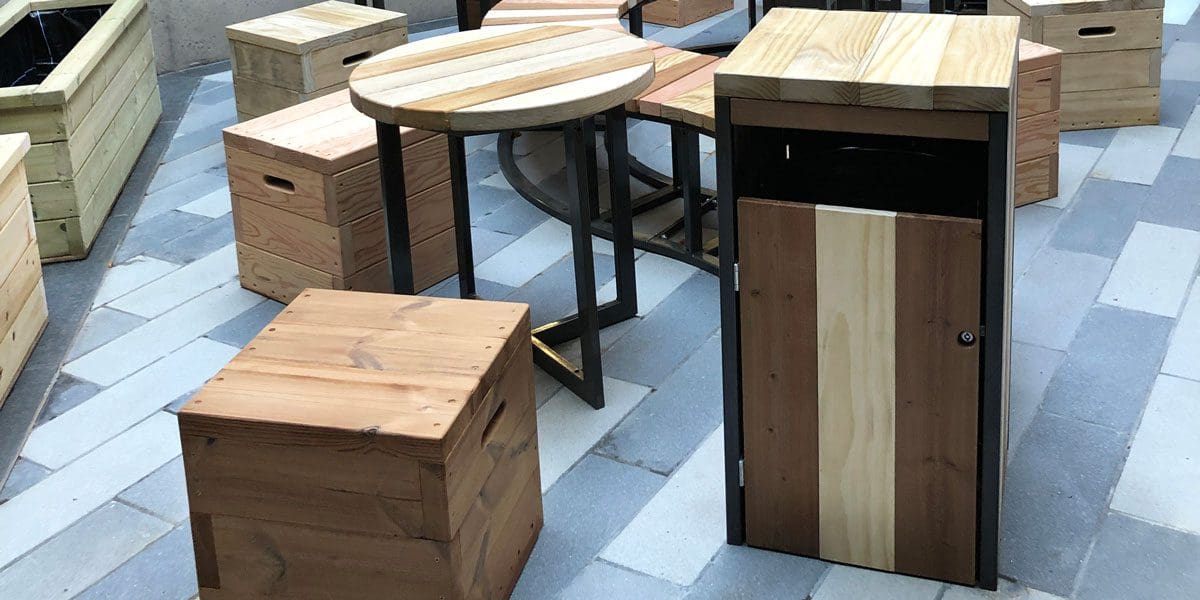 collection of varying toned wooden furniture including S shaped bench with black metal legs, circular tables with black metal legs and cube wooden single seats with handles cut out and wooden and metal bin