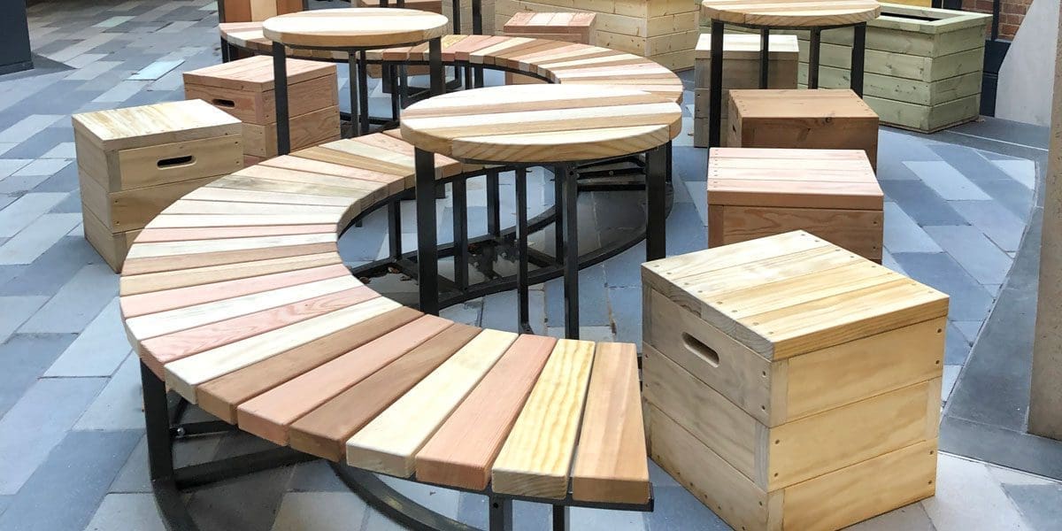 collection of varying toned wooden furniture including S shaped bench with black metal legs, circular tables with black metal legs and cube wooden single seats with handles cut out