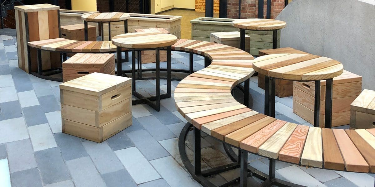 collection of varying toned wooden furniture including S shaped bench with black metal legs, circular tables with black metal legs and cube wooden single seats with handles cut out