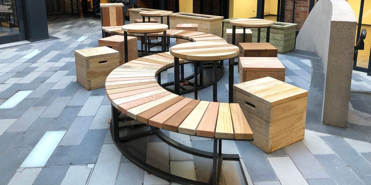 collection of varying toned wooden furniture including S shaped bench with black metal legs, circular tables with black metal legs and cube wooden single seats with handles cut out