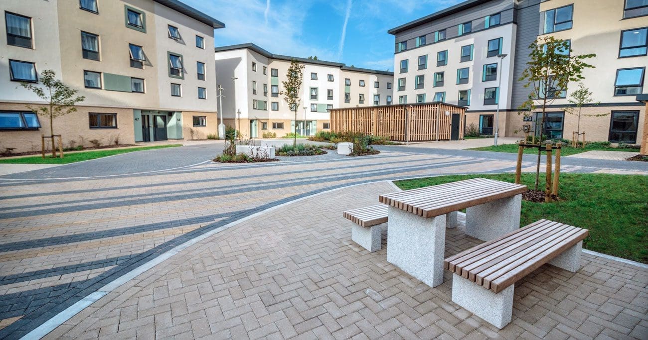 University exterior showing large cycle storage structures infront of tall buildings and wooden and concrete outdoor table and matching bench
