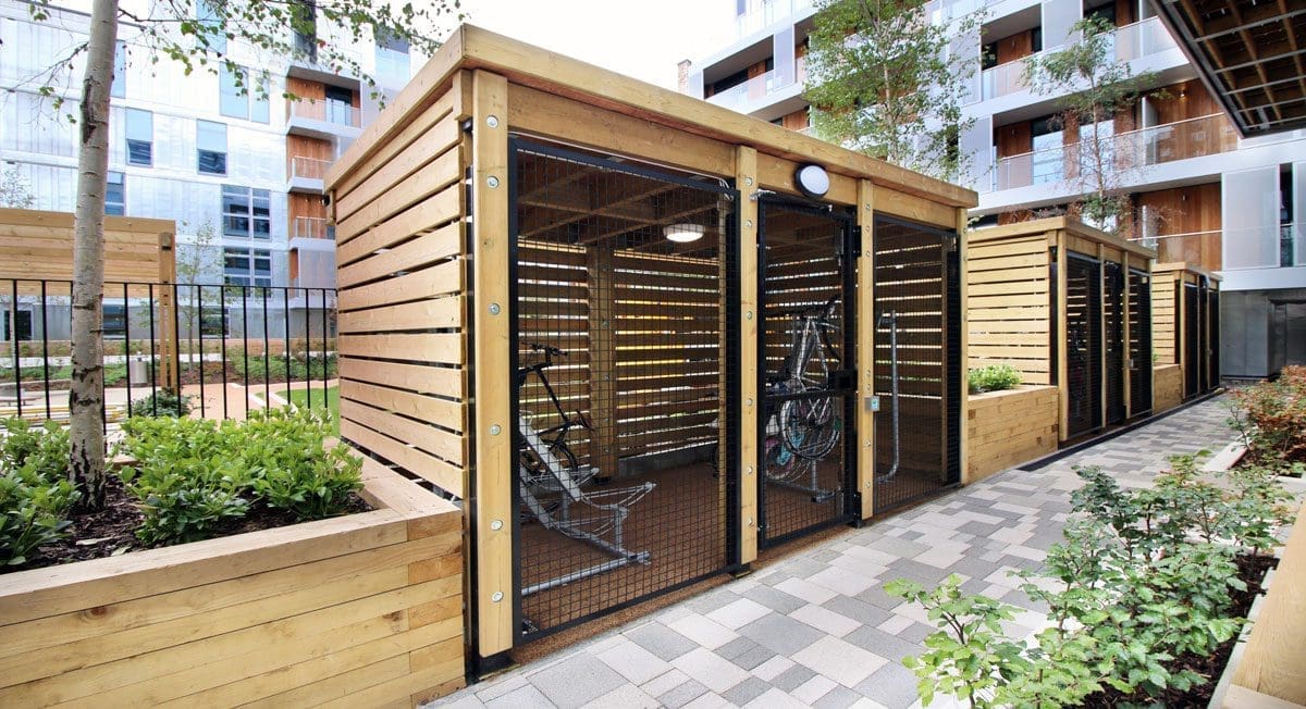 Row of wooden bicycle stores with black cage doors and metal racks inside separated with raised wooden planters