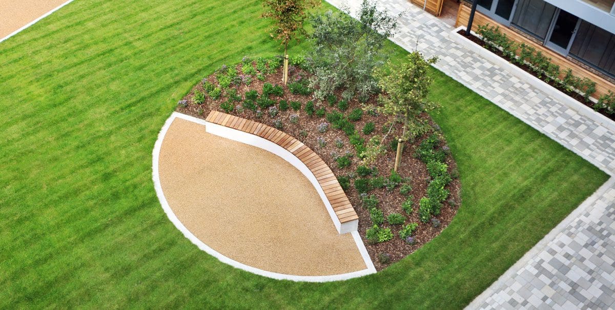 Circular landscaped ying yang symbl on grass split in the middle, half tarmic with a curved concrete bench with wooden top and half bushes and trees