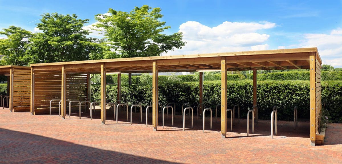 Large wooden pergola covering metal bicycle rack hoops in the ground with small thin raised planters along the left and right edges