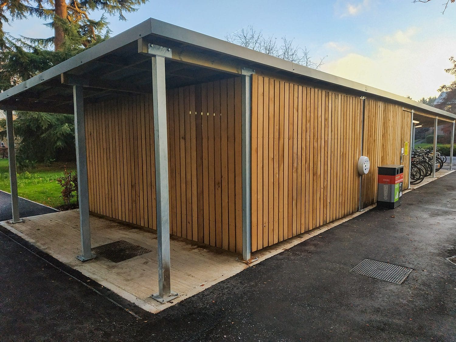 Rows of metal bike secure hoops under wooden and metal canopy connected to outdoor wooden storage area