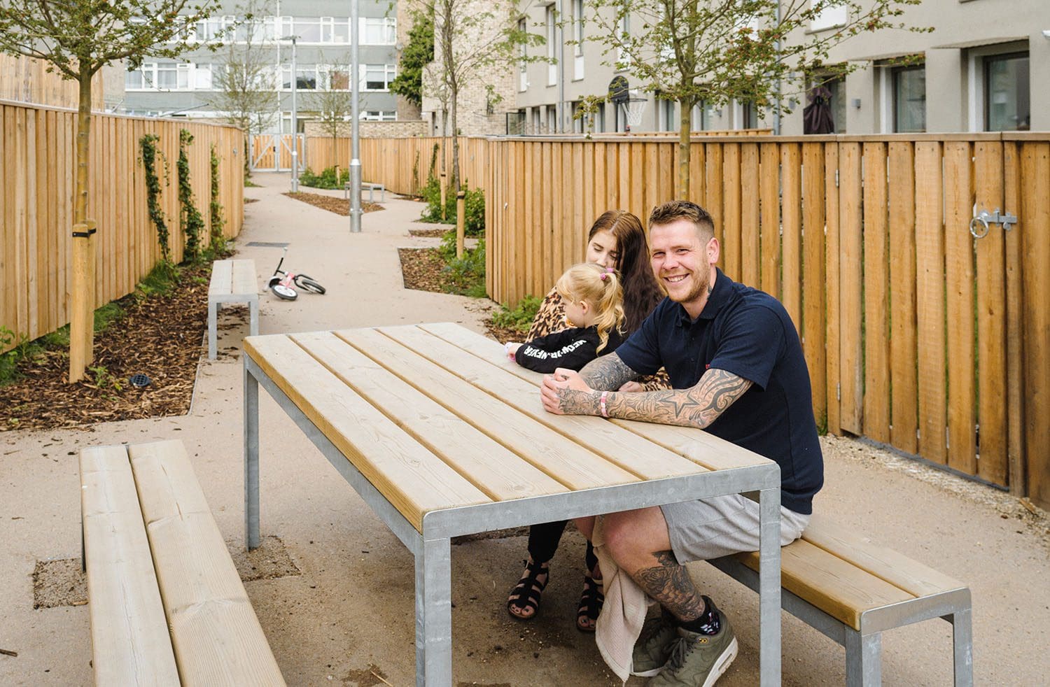 Outdoor wooden picnic table and matching benches with metal legs, happy family sat on one side smiling at the camera