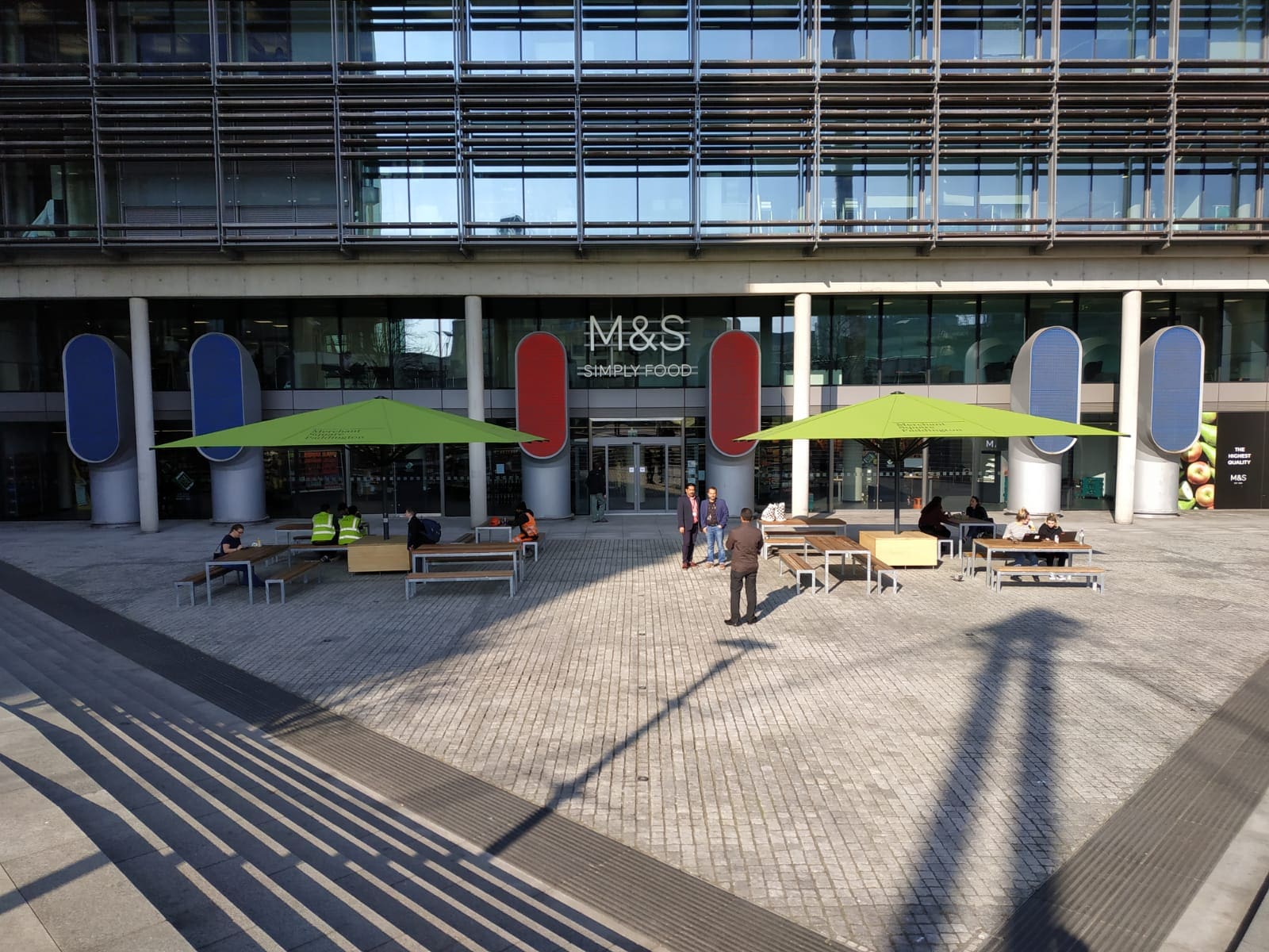 Exterior of M&S building with wooden and metal picnic benches and tables under large square green parasols