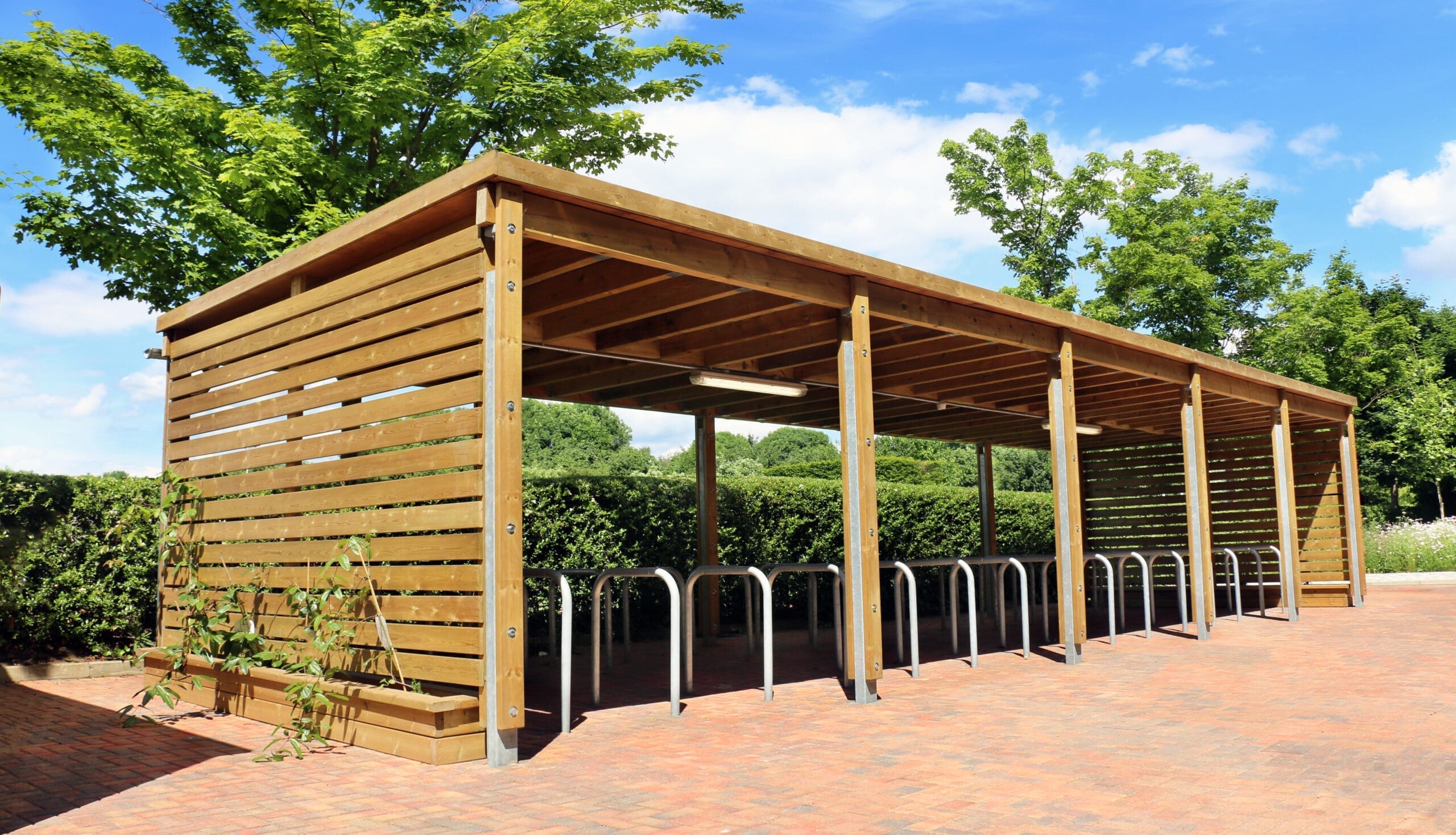 Wooden covered bike rack, with trees behind and a blue sky with clouds.