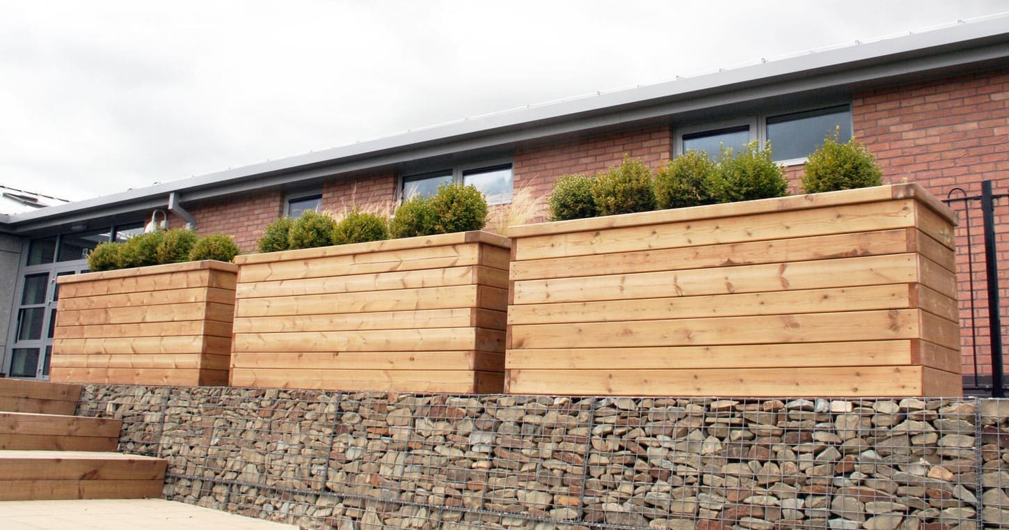 3 wooden planters filled with green bushes sat on top of a rock wall.