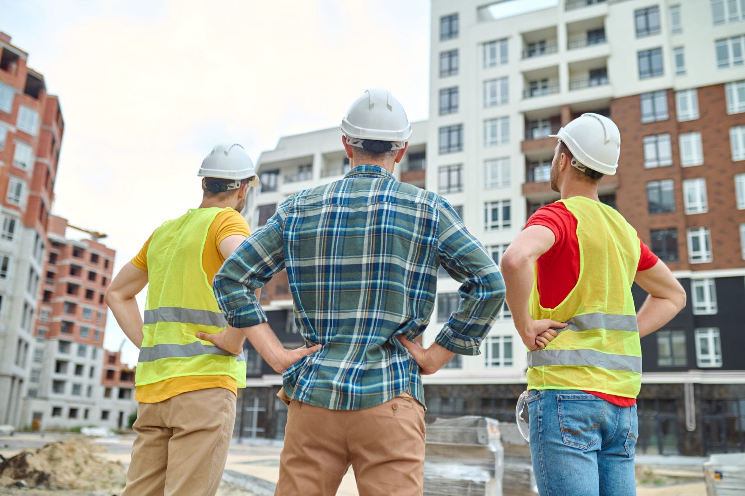 3 men stood with their backs away from the camera, each man is wearing a white protective hat. 2 of the men are wearing a high-vis jacket. The men are looking at a group of high-rise buildings.