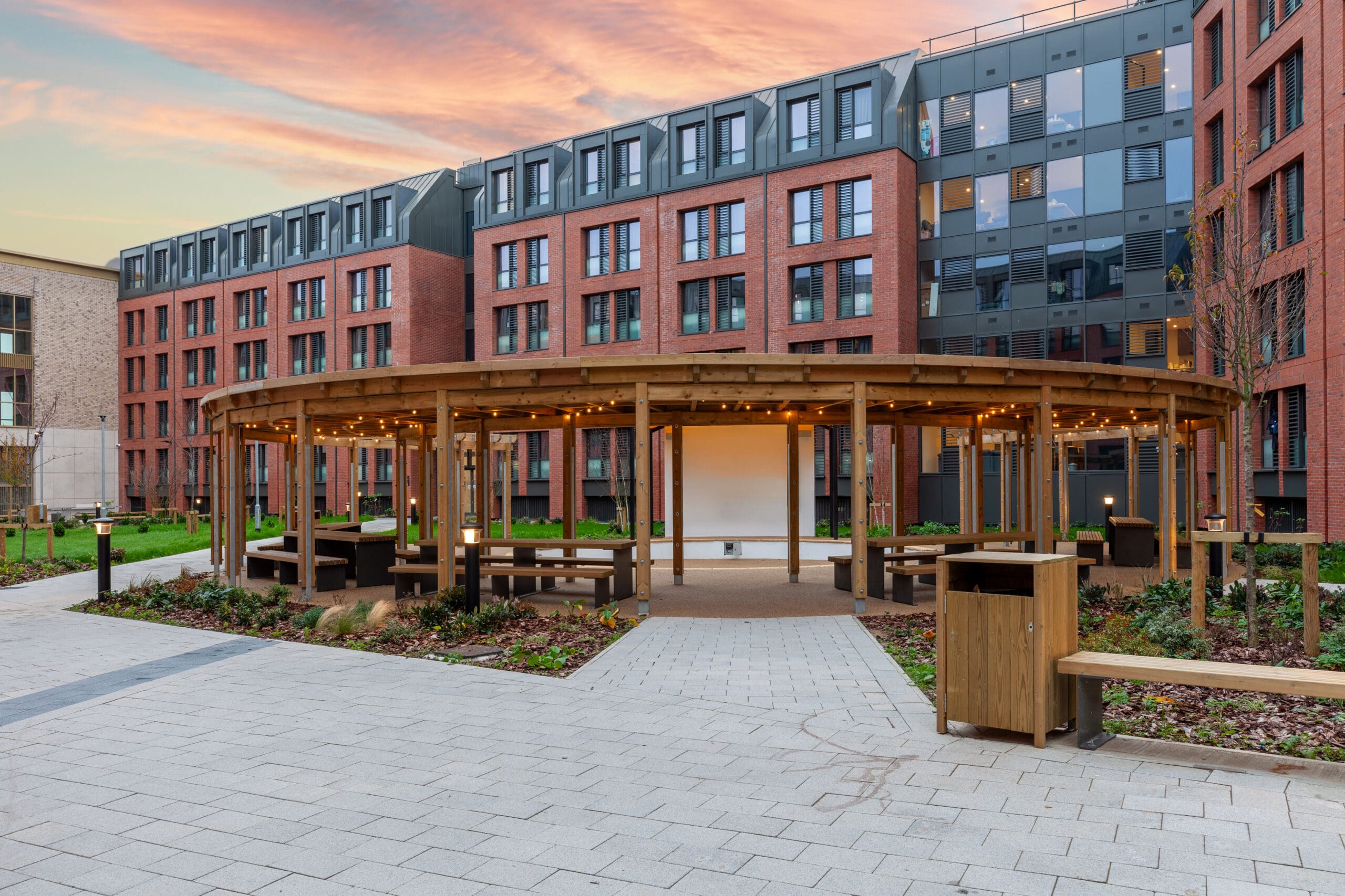 Modern building, with the sun setting in the background. Wooden canopy with benches and fairy lights. 