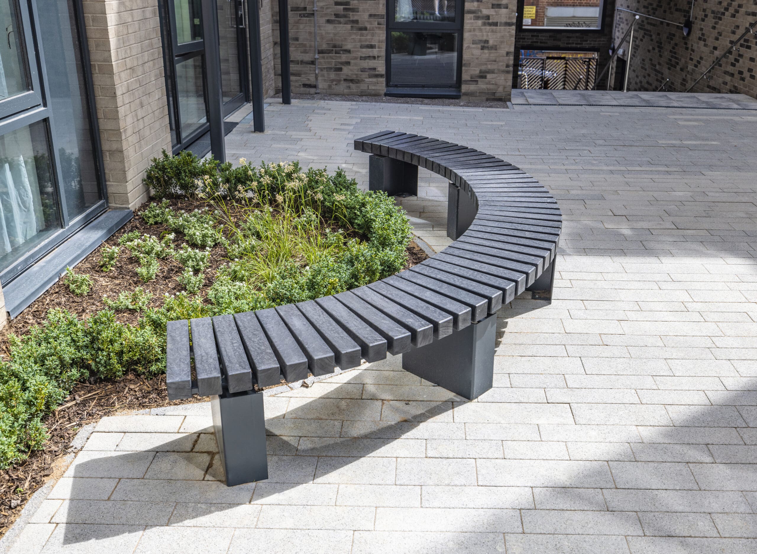 Grey curved bench, surrounded by a garden with growing flowers/shrubs. 
