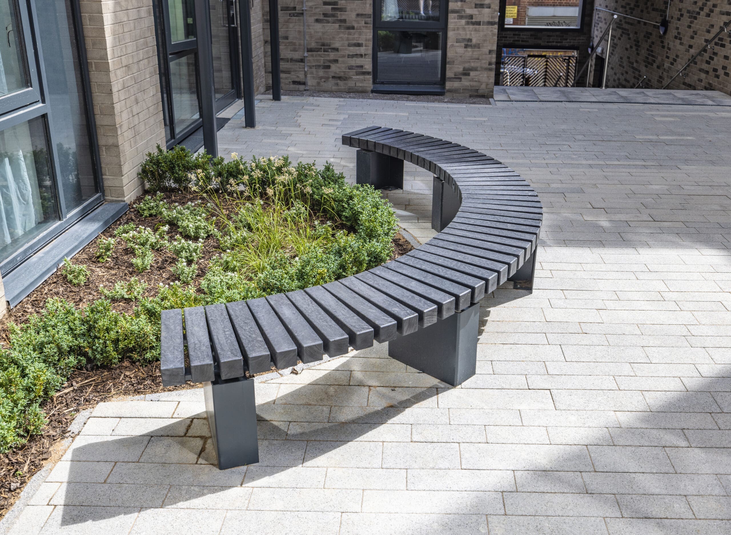 Grey curved bench, surrounded by a garden with growing flowers/shrubs.