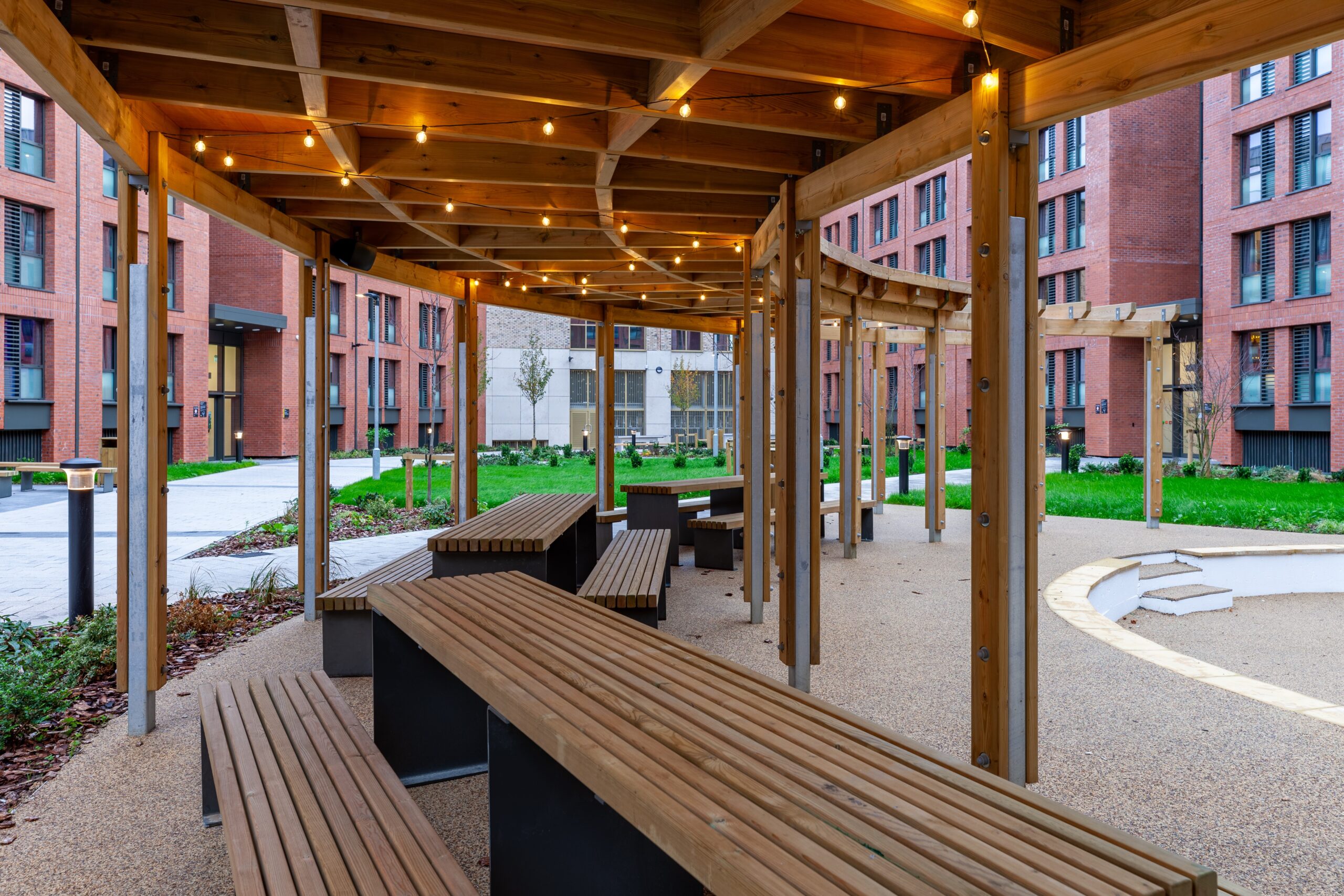 Picnic tables arranged under a curved pergola with fairy lights at, creating a cozy and inviting outdoor space.